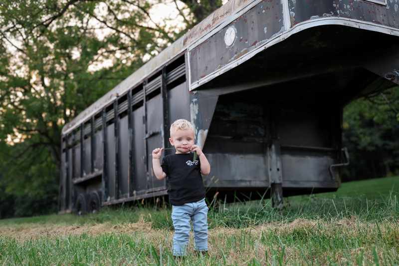 Young boy standing in a field in front of a rustic livestock trailer