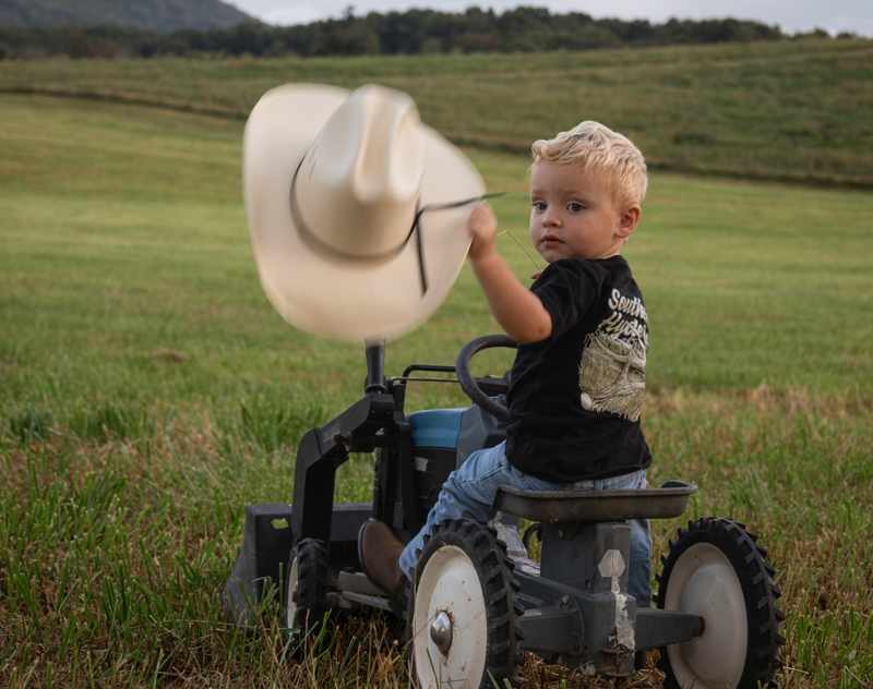 Young boy lifting his cowboy hat while sitting on a small tractor