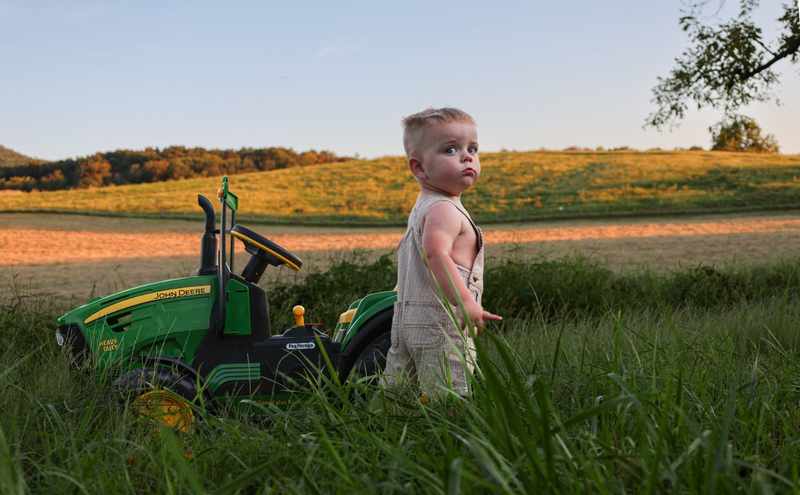 Young boy in overalls standing beside a green John Deere toy tractor at golden hour
