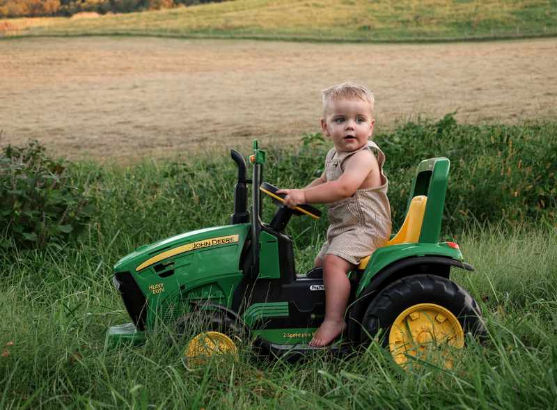 Young boy in overalls sitting on a green John Deere toy tractor
