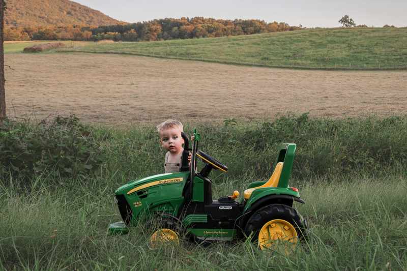 Young boy peeking over a green John Deere toy tractor in a country field
