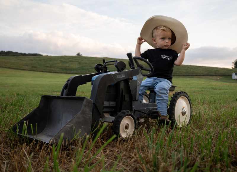 Young boy holding his cowboy hat while sitting on a small tractor under an open sky